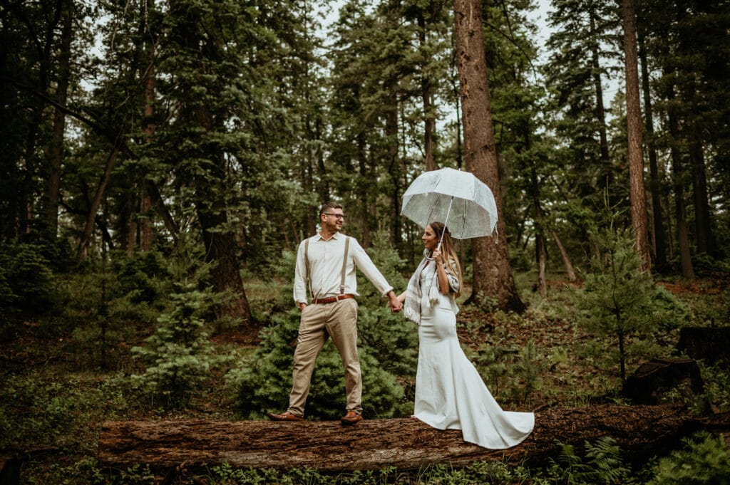 Groom leading bride in the rain as they step on a log in the forest in Arizona