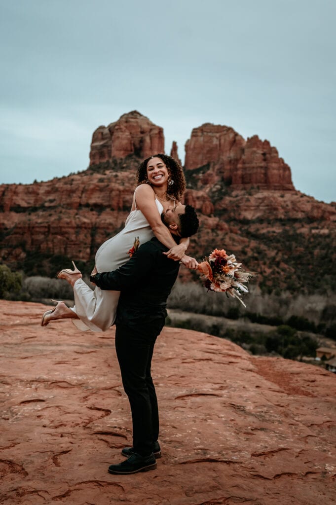 Groom lifts laughing bride on their winter elopement with Cathedral Rock in the distance
