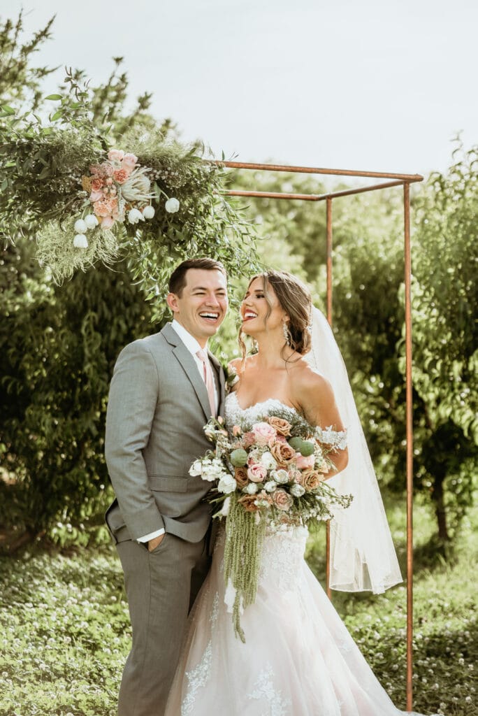Bride and groom laughing surrounded by lush greenery and overflowing floral arrangements