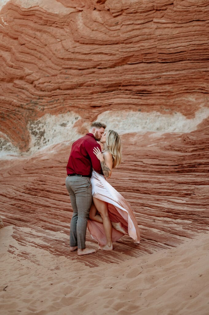 Engaged couple embrace in a kiss as they stand barefoot at their Vermillion Cliffs engagement Session