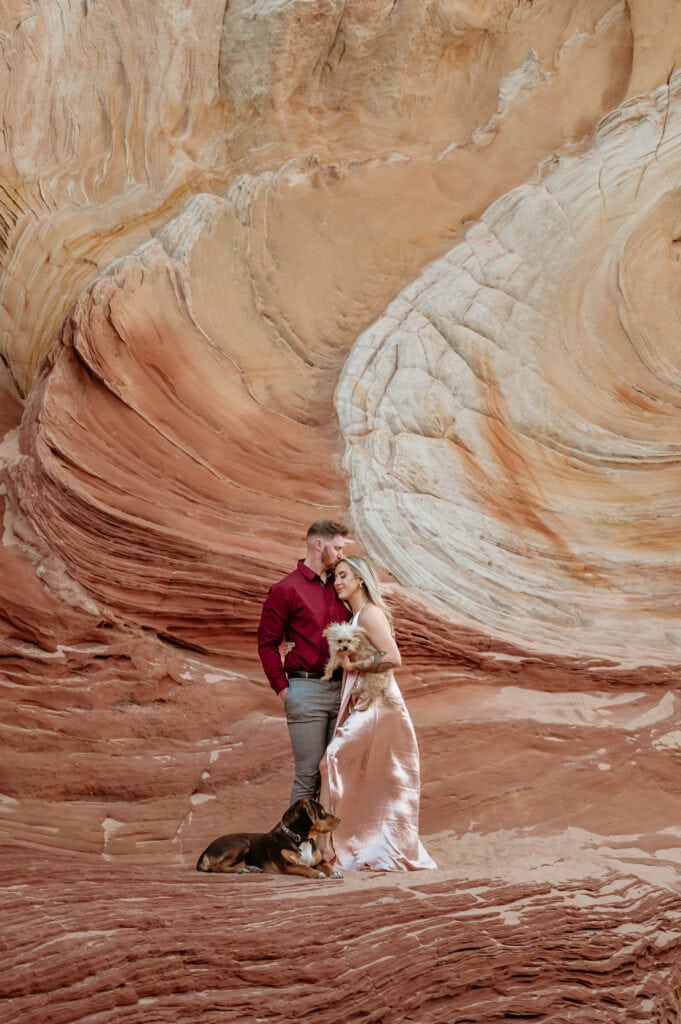 Couple embrace snuggled in with their two pups framed against a giant swirl of sandstone at their Vermillion Cliffs engagement Session