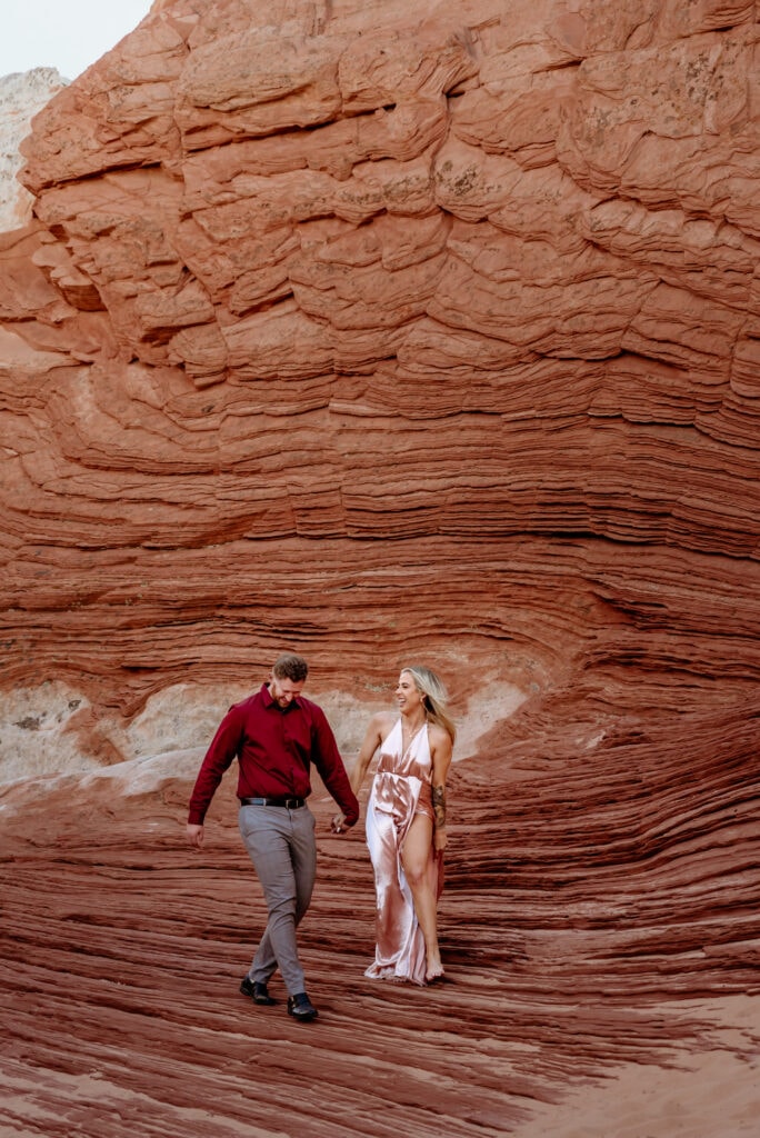 Engaged couple walking along sandstone walkway, she is wearing a shiny pink dress, and he is wearing a pair of grey slacks with maroon button up