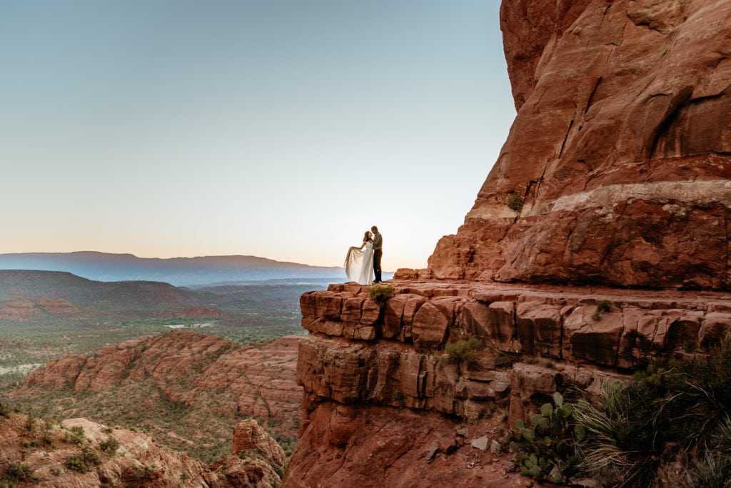 Elopement couple hold each other as the bride's dress blows in the wind at the peak of Cathedral Rock in Sedona, Arizona