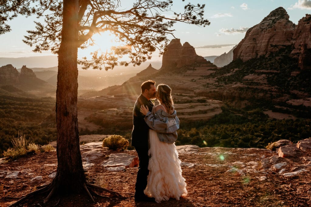 Bride and groom kiss under a tree during sunset overlooking the red rocks of Sedona below