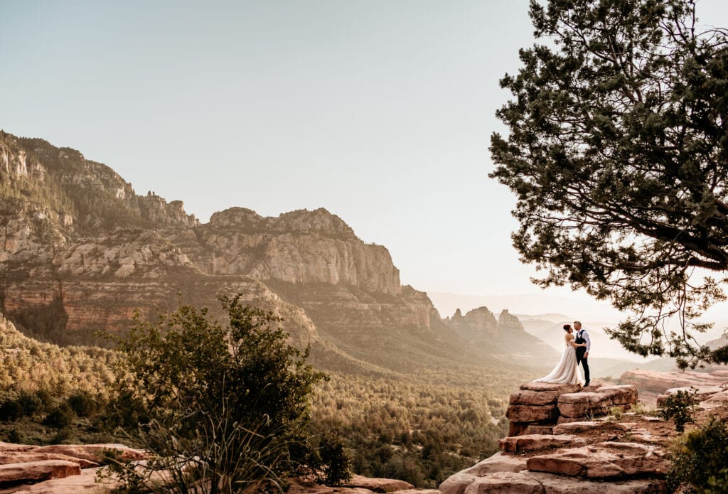Bride and groom framed by juniper trees as they stand perched on a red rock ledge overlooking the Sedona sunset during their wedding portraits